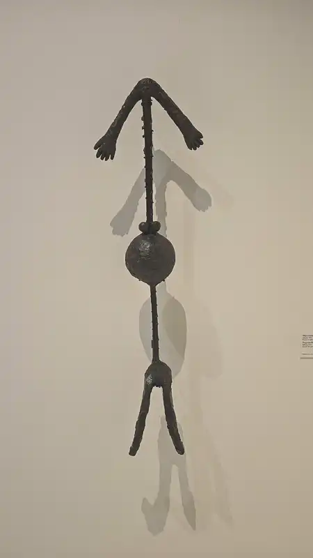 Długa, metalowa (prawdopodobnie żelazna lub brązowa) rzeźba w formie niemalże piktogramu lub ludzika z patyków. Składa się z prostej pionowej linii (korpusu), z kulą w środku, dwoma nogami na dole i ramionami uniesionymi do góry, zakończonymi dłońmi. Rzeźba jest podwieszona lub przymocowana do białej ściany i rzuca duży cień.