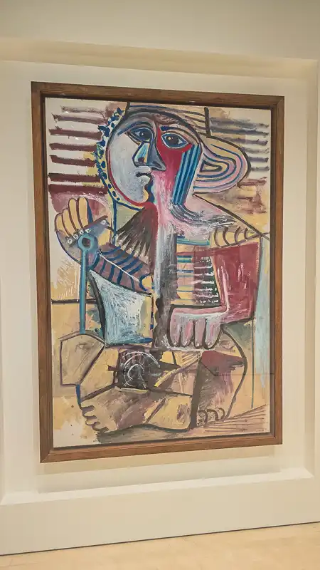 Obraz przedstawiający siedzącą postać o zgeometryzowanej i podzielonej twarzy. Lewa strona twarzy jest biała i niebieska, prawa czerwono-niebieska. Dominują żywe, choć przygaszone, barwy i mocny, linearny rysunek. W dłoni postać trzyma jakiś niebieski przedmiot (być może fajkę lub laseczkę).