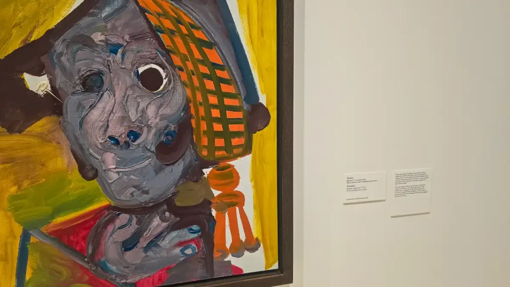 Zblliżenie na fragment ekspresjonistycznego portretu, w którym dominują bardzo grube warstwy farby (impasto) w odcieniach szarości, błękitu i pomarańczowo-żółtym tle. Postać ma przerysowane, niemal groteskowe rysy twarzy. Na ścianie obok widoczna jest etykieta z tytułem/opisem obrazu (częściowo czytelna: Sienno - po hiszpańsku i angielsku, prawdopodobnie portret Sylvette David).