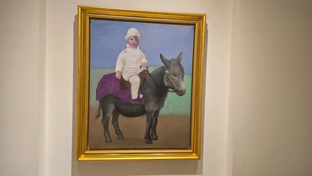 Obraz w stylu realistycznym (klasycystycznym) przedstawiający małe dziecko ubrane na biało (w czapeczce i kombinezonie) siedzące na małym, ciemnym osiołku przykrytym fioletowym kocem. Tło jest stonowane, w odcieniach zieleni i nieba.