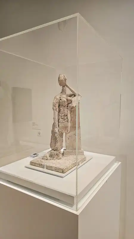 Rzeźba/model w kolorze białym lub jasnobeżowym (może z gipsu lub terakoty) przedstawiająca zdeformowaną, stojącą postać ludzką z elementami kolumnowymi lub architektonicznymi. Eksponowana w przezroczystej gablocie.