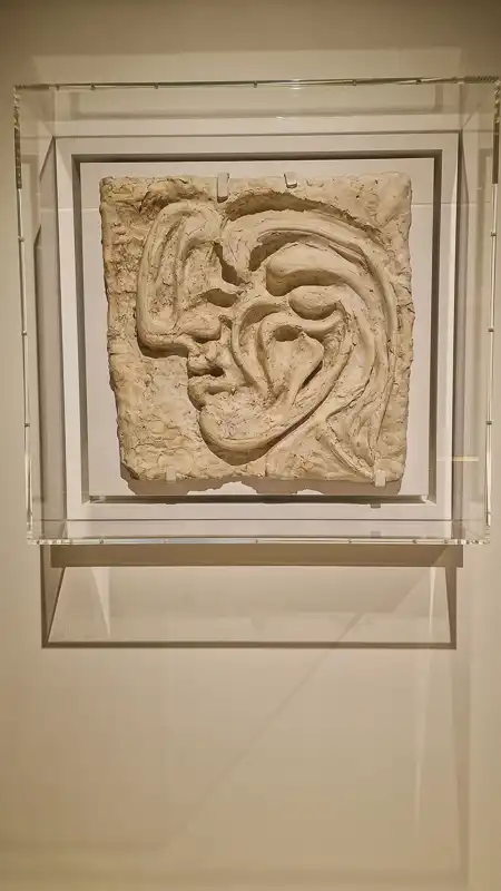 Jasna, beżowa płaskorzeźba przedstawiająca zgeometryzowany i pofalowany profil twarzy (lub dwóch nachodzących na siebie twarzy) w stylu organicznego surrealizmu. Eksponowana w przezroczystej gablocie.