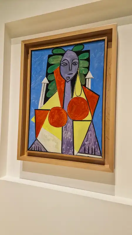 Obraz przedstawiający siedzącą kobietę w jasnych, jaskrawych kolorach (błękit, żółty, czerwony, fiolet). Postać jest mocno zgeometryzowana, o fioletowej twarzy i bujnych zielonych włosach. Na piersi widoczne są dwa duże, pomarańczowe okręgi.