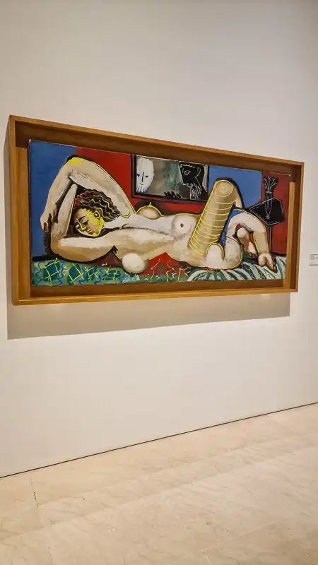 Długi, poziomy obraz przedstawiający nagą postać kobiecą leżącą na dywanie o geometrycznych wzorach. Postać ma jasną karnację i żółtą twarz. W tle widoczne są abstrakcyjne, ciemniejsze formy.