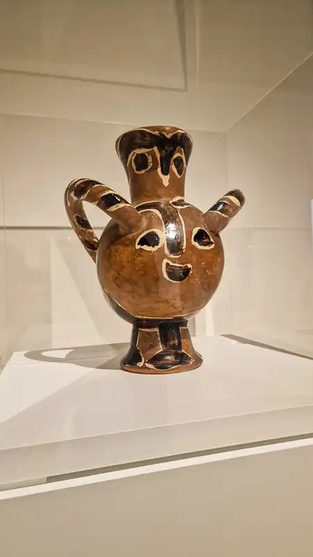 Ceramiczne naczynie w kształcie dużej, brązowej kuli z krótką nóżką i dwoma uchwytami, które wyglądają jak uszy lub ramiona. Na korpusie narysowany jest uśmiechnięty, uproszczony pyszczek w stylu stylizowanego słońca lub maski.